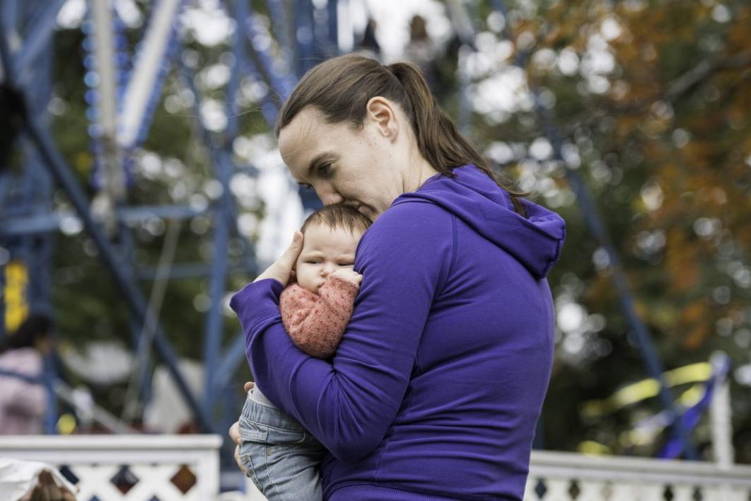 A young woman holding a baby in front of a ferris wheel at the University of Rochester's Meliora Weekend